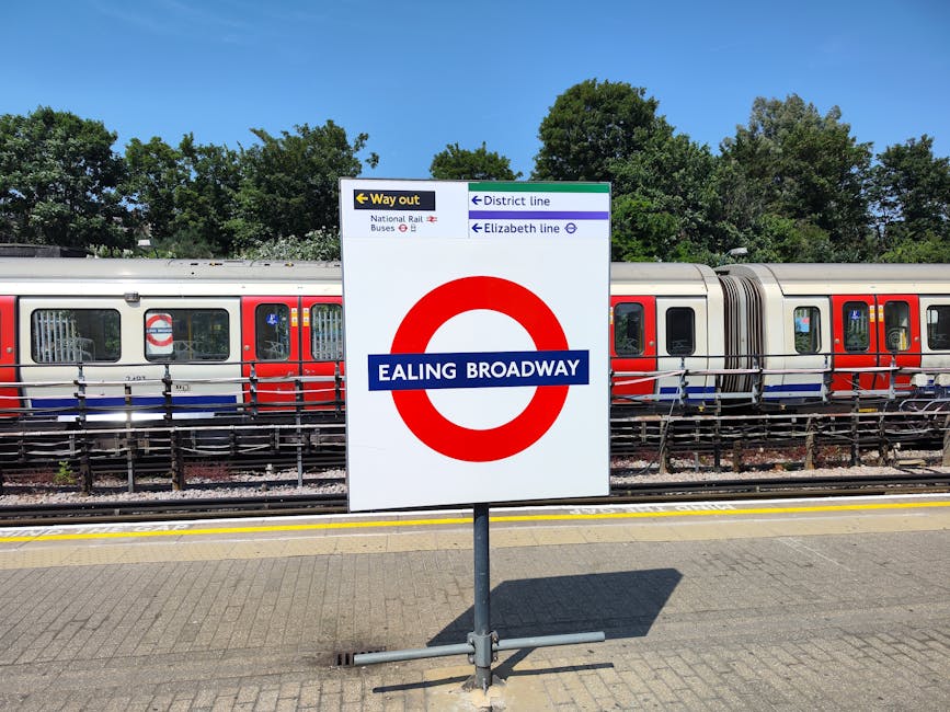 A close-up view of a white station sign with a red circle and blue horizontal bar displaying 'EALING BROADWAY', situated on a paved train platform near Barkingside. Behind the sign, a modern London Un