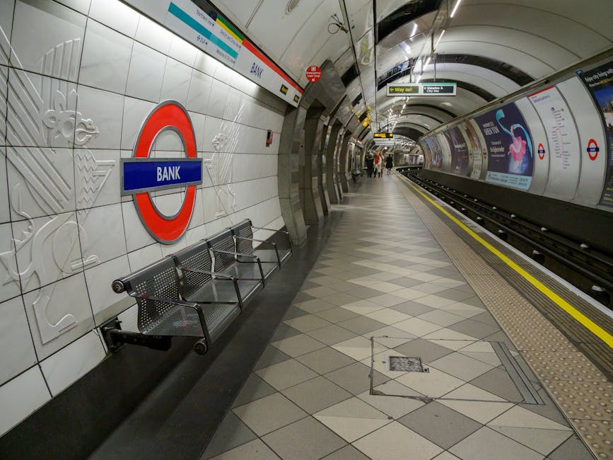 A close-up view of a white station sign with a red circle and blue horizontal bar displaying 'EALING BROADWAY', situated on a paved train platform near Barkingside. Behind the sign, a modern London Un