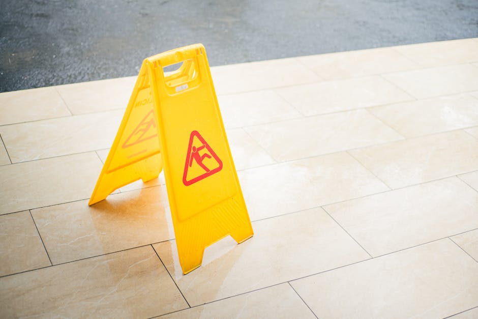 A bright yellow, portable wet floor warning sign with a red triangle and a black icon of a person slipping, placed on a smooth, light-colored tiled walkway outside a building. The tiles have a glossy 